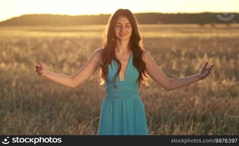 Attractive brunette woman in stylish blue dress with arms outstretched spinning around in the glow of a beautiful sunset. Young female enjoying nature, sunlight, freedom as she whirls around with eyes closed at sunset against a golden wheat field background
