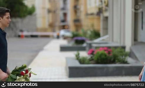 Attractive brunette woman receiving bunch of flowers on romantic date. Handsome young guy giving bouquet of beautiful flowers to his pretty girlfriend over cityscape background. Side view.