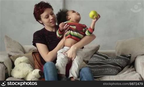 Attractive Caucasian mother and her curly mixed-race toddler boy sitting on the sofa and playing. Loving mother and adorable little son with apple in his hand spending leisure and having fun at home. Slow motion.