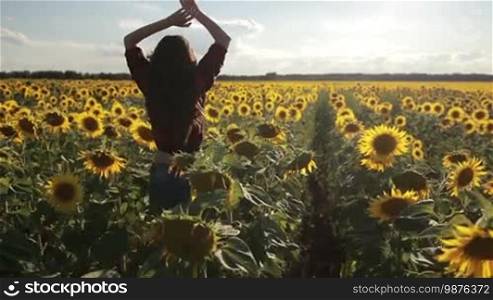 Attractive hipster female in stylish clothing enjoying leisure and freedom while running through blooming sunflower field with raised arms over amazing landscape. Back view. Charming woman delighted with sunflowers on sunny summer day. Slo mo.