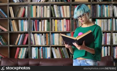Attractive hipster student with blue hair in stylish eyeglasses reading a book in the library. Portrait of a serious female college student reading a book over the library's bookshelves background.