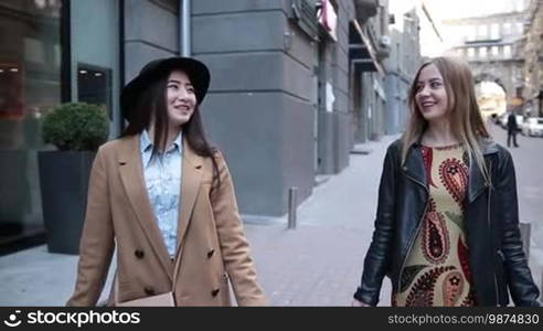 Attractive teenage girlfriends walking with shopping bags on the street, looking happy. Stylish Asian woman in black hat chatting with pretty Caucasian female friend while strolling down the sidewalk after shopping. Slow motion.