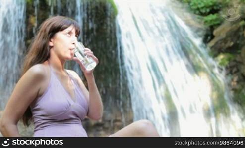 Attractive woman drinking water from plastic bottle by the waterfall