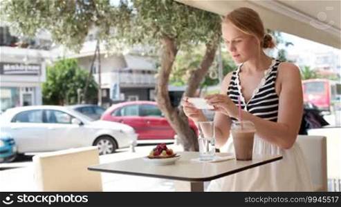 Attractive woman taking picture of a pastry on her smart phone as she sits at a table at an open-air restaurant enjoying refreshments