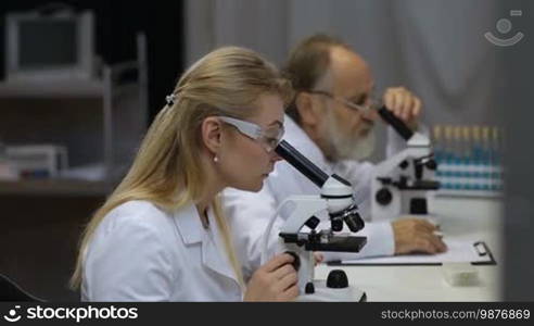 Attractive young female scientist and her senior male supervisor pipetting and microscoping in the life science research laboratory. Young female research scientist and senior male supervisor preparing and analyzing microscope slides in research lab.