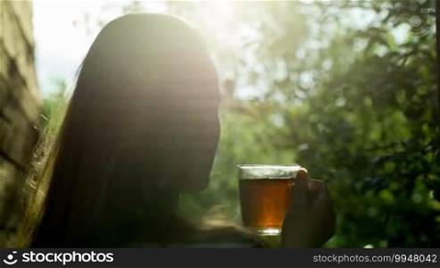Back shot of a young woman having hot tea at sunset. She is relaxing and enjoying a nature scene with green trees and the evening sun