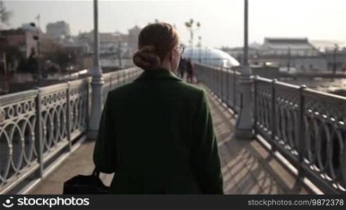 Back view of a beautiful woman walking on an iron bridge over a cityscape background. Rear view of a businesswoman strolling alone on a pedestrian bridge in the city. Slow motion.
