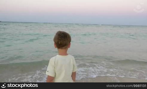 Back view of a little boy alone on the beach. He is standing by the sea and looking as waves come close to his feet