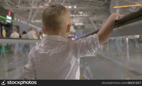 Back view of a little boy moving through the crowded shopping mall on flat escalator. People traffic from both sides of him