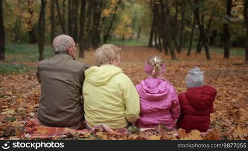 Back view of a positive multi-generation family embracing each other while sitting on yellow fallen leaves in the park over a colorful autumn background. Grandparents and grandchildren spending time together in the fresh air as they walk in the park in the fall.