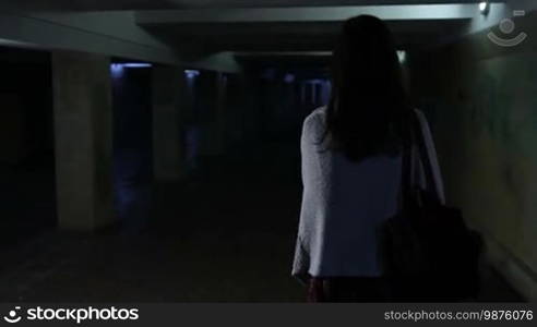 Back view of an attractive woman, scared by a stranger, running through a dark underground passage. Frightened young girl running away from a stranger through an underpass at night. Slow motion. Steadicam stabilized shot.