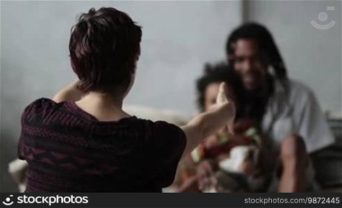 Back view of Caucasian mother sitting on the floor with open arms, waiting for mixed-race cute child to embrace. Blurry African American father with dreadlocks sitting on sofa and holding little boy. Cute kid runs away from mom's embrace. Slow motion