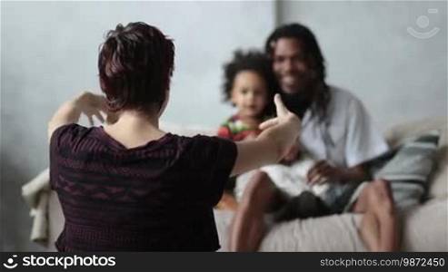 Back view of Caucasian mother with open arms sitting on the floor to embrace her cute mixed-race toddler son over blurry African American father sitting on the sofa with little child background. Slow motion.