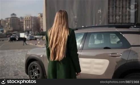 Back view of fashionable female driver with long blonde hair wearing emerald green coat opening car's door and getting in the automobile. Rear view of attractive woman getting into parked car in daylight over urbanscape background. Slow motion.