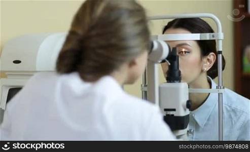 Back view of professional oculist checking woman vision with modern medical equipment in ophthalmologist's office. Female optometrist examining beautiful woman's eyes with slit lamp in eye clinic.