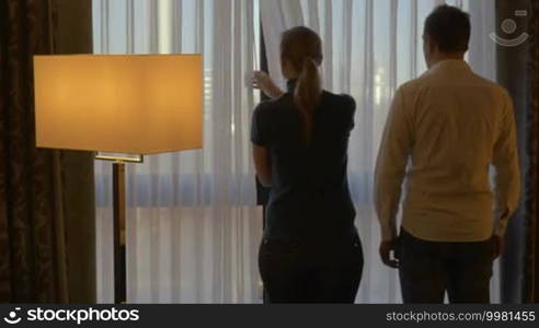 Back view of romantic couple at home or in a hotel room. Woman opening the curtain, they embracing gently and looking out the window. Happy time together