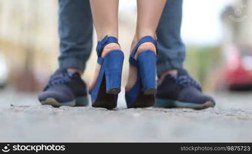 Back view of woman's legs in high heels standing on tiptoes to kiss her boyfriend during a romantic date. Close-up. Couple's feet standing on cobblestone street while woman raising up on tiptoes to kiss her beloved man.