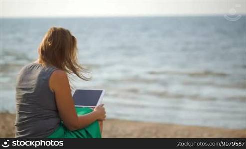 Back view of woman sitting on the beach on a windy summer day and using a tablet computer