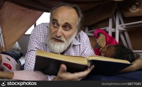 Bearded aged man and granddaughters reading a book together in cubby house made of blanket and chairs. Affectionate grandfather reading fairy tales to his adorable little mixed race girls while spending leisure at home. Dolly shot. Slo mo.