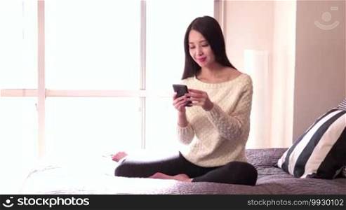 Beautiful Asian woman on bed at home, texting with mobile phone and smiling, pretty Japanese girl using smartphone for text messaging, internet and email. Happy people, technology, lifestyle, fun