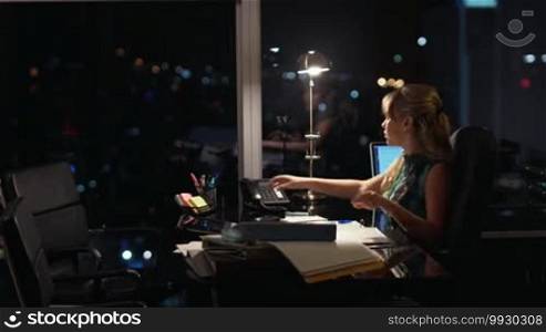 Beautiful blond business woman working overtime at night in executive office, answering phone call and talking on wired telephone. City lights are visible in background from a large window
