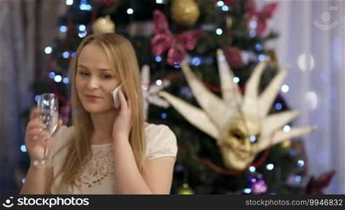 Beautiful blond young woman smiling and drinking champagne while talking on a mobile phone, with a Christmas tree decorated with lights, baubles, bows, and a Venetian mask in the background