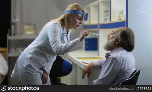 Beautiful blonde female physician in lab coat examining senior man's throat with tongue depressor at clinic. Young female ENT specialist checking elderly male patient's throat using a tongue depressor and head mirror to look inside mouth and tonsils.