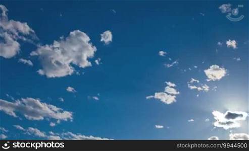 Beautiful blue sky background with fluffy white clouds and sun flare where the sun is shining out from behind a small cloud to the right of the frame