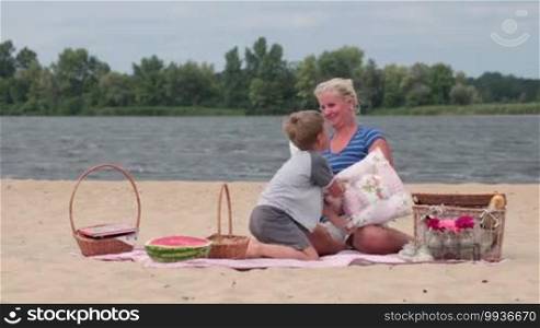 Beautiful excited mother and son having fun on a picnic on the river bank during the weekend and playing in a pillow battle.