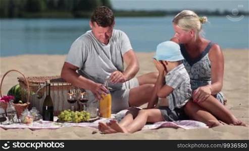 Beautiful family on summer holidays picnicking on the beach. Father pouring orange juice in glass and son drinking it