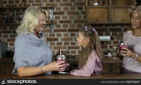Beautiful grandmother with mason jar of berry smoothie chatting with lovely little granddaughter. Three generation family having breakfast in the kitchen. Cute girl hugging her charming mother after mom brings smoothie jar to her girl.