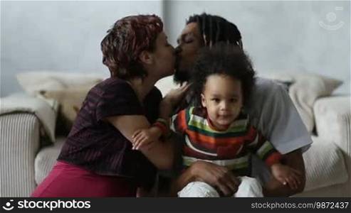 Beautiful mixed couple in loving embrace and sensual kiss. Loving mixed race parents with adorable mixed race child sitting on the floor and enjoying weekend at home. Caucasian mother with eyes closed kissing her African American husband with passion.
