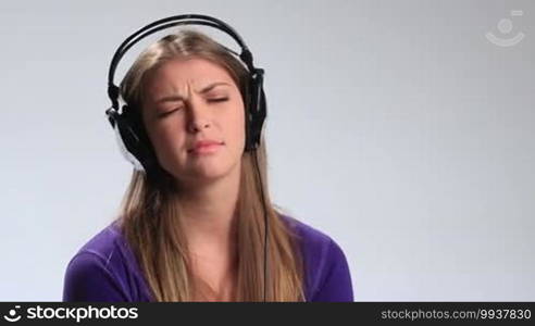 Beautiful sad pensive young woman in headphones listening to music on white. Attractive brunette girl moving slowly to music, being gloomy and thoughtful, looking at the camera with eyes full of sadness, closing her eyes, and putting her hand on headphones.