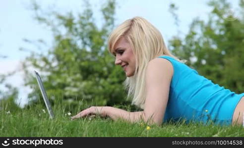 Beautiful smiling blonde woman lying in the grass working with a laptop computer