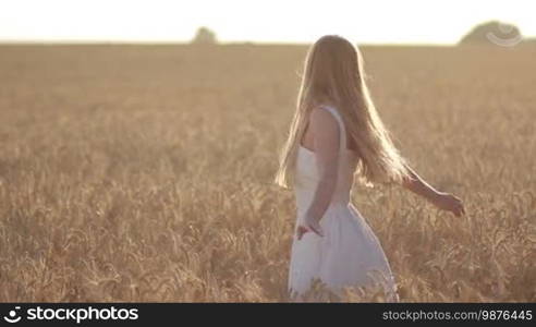Beautiful smiling woman with flying long blond hair spinning around in ripened wheat field in rays of setting sun in summer. Cheerful girl in white dress relaxing outdoors at sunset during holidays. Slow motion.