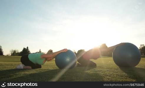 Beautiful sporty senior females in sportswear training and stretching with fitness balls on green grass in park in glow of sunset. Adult fit women exercising with sport balls outdoors, toning arms and abdominal muscles. Dolly shot.