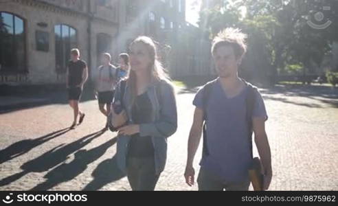 Beautiful student couple walking on university campus, holding books and chatting. Joyful hipster college friends going to lesson through park outside university building and talking with group of classmates in the background. Slow motion. Stabilized shot