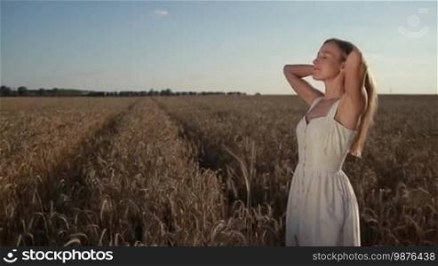 Beautiful young lady in summer dress playing with her long blond hair as she stands in golden wheat field in sunlight. Charming female posing in cereal field over blue sky background. Slow motion. Steadicam stabilized shot.