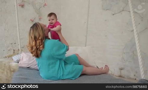 Beautiful young woman sitting on bed in stylish bedroom, bonding with her adorable baby girl while family spending leisure at home. Loving mother gently kissing her infant child. Slow motion. Steadicam stabilized shot.