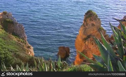 Blick auf Felsengebilde / Stein im blauen Meer von einem grün bewachsenen Felsen; die Sonne spiegelt sich im Wasser. Küste der Algarve, Portugal.