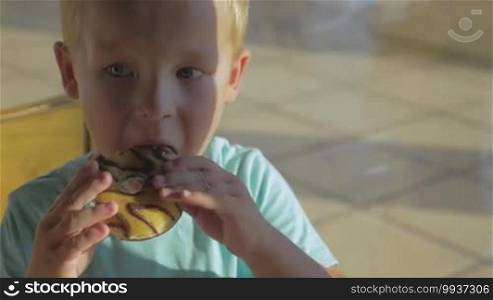Blond boy biting chocolate donut and drinking water from clear plastic glass close up view with crumbs on lips
