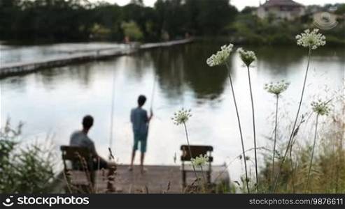 Blurry teenage son and father with fishing rods angling on wooden pier at tranquil pond. Foreground wildflowers swaying in the wind. Happy family spending leisure fishing at freshwater pond on summer day.