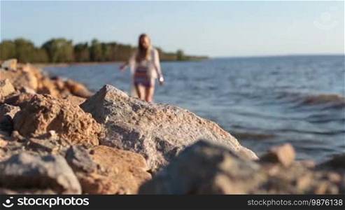 Blurry young fashionable blonde female in mini skirt walking barefoot along seaside. Focus on rocky coastline. Attractive woman enjoying summer vacation, strolling on the beach.