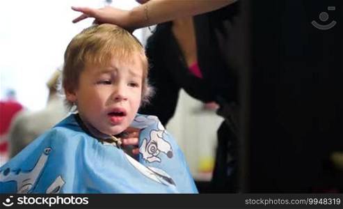 Boy unwilling to wear a cape and taking it off, while hairdresser cutting his hair
