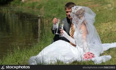 Bride and groom drink champagne sitting on the shore of Lake