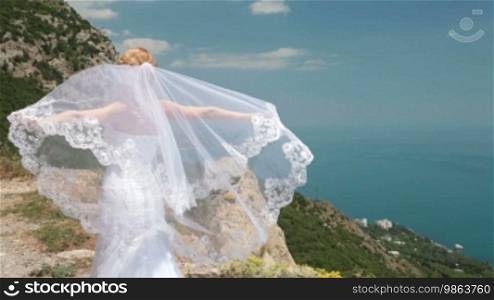 Bride stands on cliff with a flowing veil in the wind against the blue sea and sky