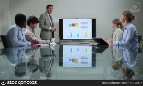 Business people talking during corporate presentation in meeting room, with charts and analytics in background on TV. 11 of 20