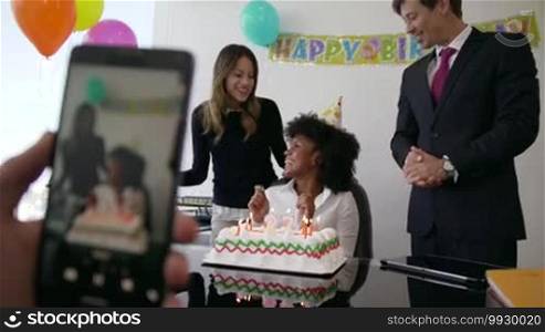 Business woman celebrating her birthday and doing a party with colleagues in her office. A friend records a video on a mobile phone of her blowing out candles on a cake. Medium shot