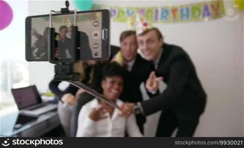 Business woman celebrating her birthday and doing a party with colleagues in her office. A friend holds his phone with a selfie stick and takes pictures of his fellow coworkers smiling. Medium shot
