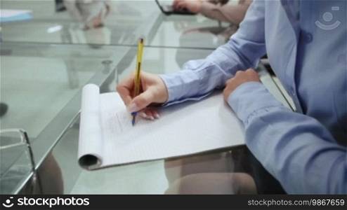 Business woman writing during corporate presentation in meeting room, with colleague out of focus in background, 3 of 20